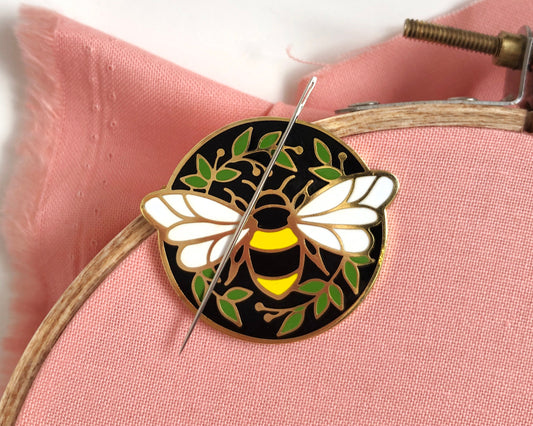 image shows a small metal needleminder. the design is a bee outlined in gold, against a black background. a needle rests against the top of the bee. 